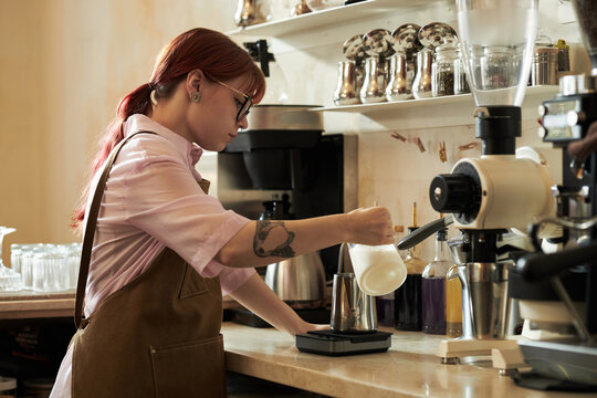 Young adult Caucasian woman with red hair preparing coffee using milk frother and scale in cafe, wearing apron and glasses, standing at counter with professional coffee equipment