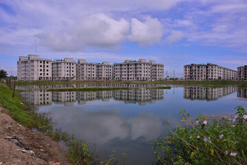 Modern residential apartment buildings reflected in a calm pond, with blooming wildflowers and greenery in the foreground. A scenic blend of urban development and natural beauty in Bangladesh