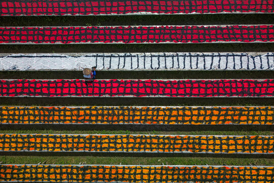 Aerial view of vibrant textiles laid out in neat rows, creating a striking mosaic of color, with a lone figure tending to the fabrics, Mojolaban, Central Java, Indonesia.