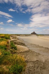 Scenic view of Mont Saint-Michel in Normandy, France, on a sunny day with dramatic clouds, tidal flats, and green grass along the walking path.