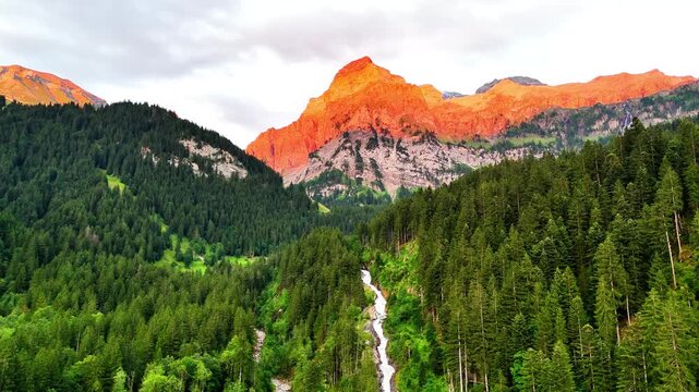 A vibrant mountain scene in Lenk, Simmenf&auml;lle, Switzerland, with a glowing orange peak above dense pine forests, a winding river, and a cascading waterfall cutting through the lush green valley