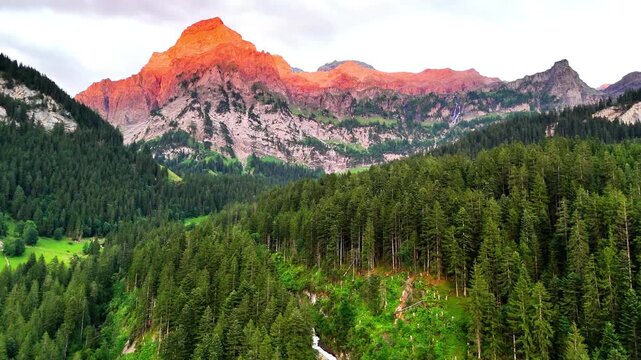 Sunrise illuminates alpine peaks above Lenk Simmenf&auml;lle, Switzerland, with vibrant orange glow, dense pine forest stretching across the valley, and a waterfall flowing through the lush green landscape