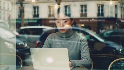 View through a cafe window of a young Asian woman focused on her laptop. City lights and car traffic are reflected on the glass, blending the quiet workspace and the busy street. - Powered by Adobe