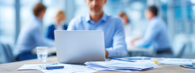 Close-up of businessman at desk with charts and laptop, blurred colleagues in background, representing corporate teamwork and data analysis.
