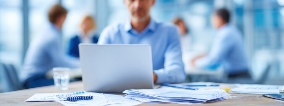 Close-up of businessman at desk with charts and laptop, blurred colleagues in background, representing corporate teamwork and data analysis.