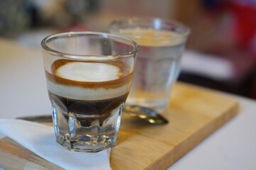 Delicious layered coffee shot with creamy milk foam served in a small glass on a wooden tray with a glass of water in a cafe