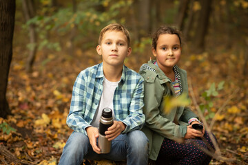 Kid teenager boy and girl hiking in the forest relax sitting on stump and drinks from thermos in autumn forest