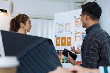 Three people are sitting at a table with a whiteboard behind them. One of the people is holding a pen and looking at a diagram on the board. Scene is focused and serious
