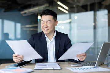 Photo of a young Asian man sitting at a desk in an office and working with documents with a smile