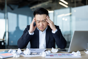 Close-up photo of a sweet Asian man sitting tiredly at his desk in the office, looking at crumpled paper, holding his head
