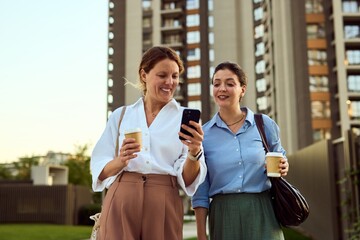 Two Businesswomen Smiling Over Phone While Holding Coffee in Urban Outdoor Setting During Daily Commute Meetup