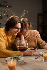 Smiling couple looking at smartphone while having dinner at home. Real woman and man sharing social media during romantic date.