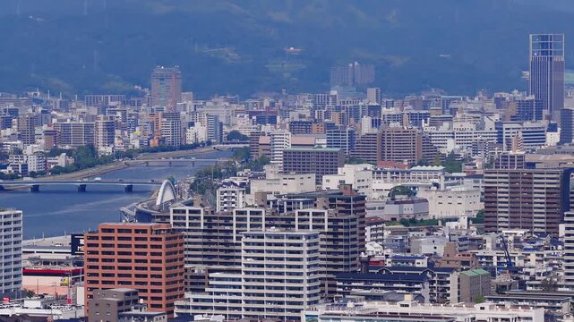 Aerial view of Hiroshima city, Japan, showing modern and traditional buildings, a river running through downtown, and green mountains in the background.