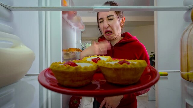 Woman opening fridge and putting spoiled cupcake inside with disgust, view from inside of fridge. Young female with repulsed and irritated expression reacting to bad taste. Lady holding dessert away