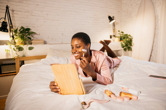 Black woman enjoying self-care facial roller routine in bed