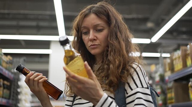 Young woman with curly hair carefully reads the label on a glass bottle of olive oil, comparing two different products before making a purchase decision in a brightly lit grocery store