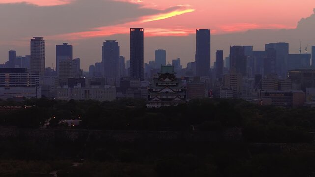 Aerial view of Osaka Castle surrounded by greenery with the city skyline in the background. The sky transitions from orange to deep hues at sunset.