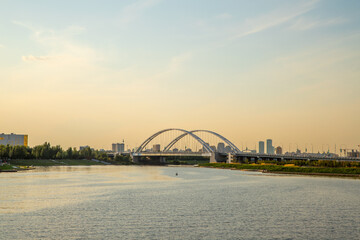 Sunset view over a river with a modern steel arch bridge and city skyline in the background. Astana, Kazakhstan, Argar Bridge over the Ishim River