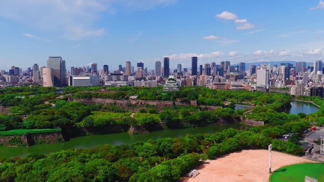 Aerial view of Osaka Castle surrounded by trees and a moat, with the modern city skyline in the background under a bright blue sky.