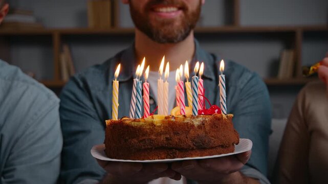 Close up unrecognizable man holding birthday cake with burning candles happy male smiling make wish near friends three unknown people at home festive party celebrating event together congratulation