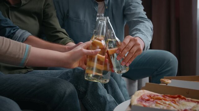 Close up four unrecognizable friends clinking bottles at home part drinking beer alcohol addiction dependency multiracial people with drinks communicating together on sofa diverse women men with pizza