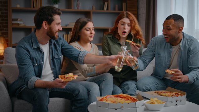 Happy multiracial four friends home party clinking bottles beer alcohol eating pizza on couch diversity buddies cheers with drinks talking communicate friendship birthday event celebration men women
