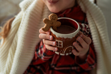 Closeup Little girl in pajamas sitting by the window drinking hot cocoa from a Christmas cup with a gingerbread man. Winter holidays.