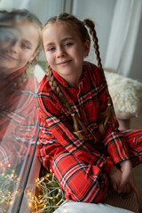 Little girl in pajamas sits by the window and waits for a gift from Santa Claus. Christmas mood. Holidays.