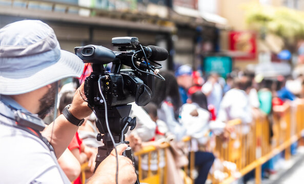 Man records a public gathering using a professional video camera mounted on a tripod during a sunny outdoor event.