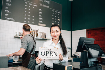 A smiling female cafe worker stands behind the counter, holding a white "OPEN" sign. A chalkboard menu is on the wall behind her, and a male colleague works nearby.