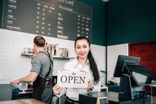 A smiling female cafe worker stands behind the counter, holding a white "OPEN" sign. A chalkboard menu is on the wall behind her, and a male colleague works nearby. - Powered by Adobe
