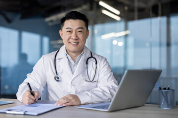 Close-up portrait of a young Asian male doctor in a white coat sitting at a desk with a laptop, making notes in a notebook