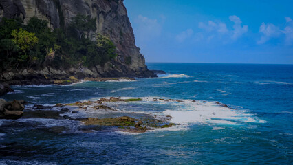 Green cliffs tower over the calm blue sea, with white waves crashing against the rocks and a clear sky stretching out.