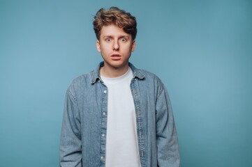 Obraz premium Young man in denim shirt looking surprised against blue background.