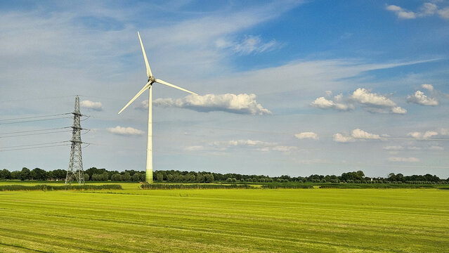 Green energy power generation via wind turbine in Dutch farming landscape with electric line and blue sky.