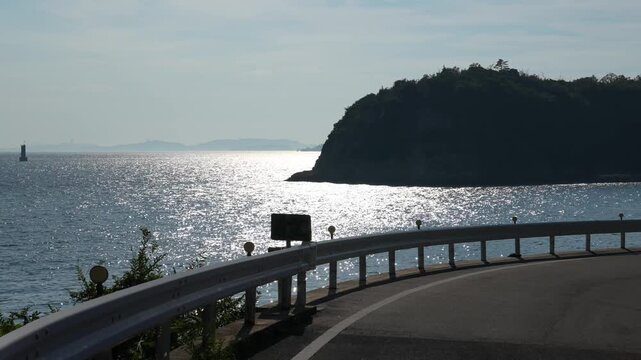 On Naoshima's winding coastal road, the Seto Inland Sea sparkles in the setting sun beyond guardrails, with lush islands and hazy mountains outlining tranquil summer coastal scenery.