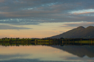 Blick über den Forggensee auf Wiesen, Weiden und Berge im letzten Sonnenlicht bei Sonnenuntergang, Bayern, Deutschland