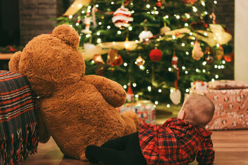 Baby with teddy bear by Christmas tree with gifts and lights in cozy holiday home