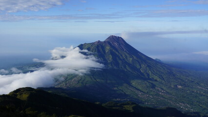 Majestic volcanic mountain peak rising above a cloud layer with lush green slopes under a clear blue sky, showcasing a grand natural landscape.