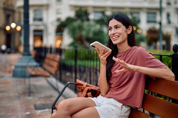 Young woman sitting on a park bench, excitedly recording a voice message on her smartphone in a...