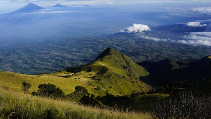 Expansive aerial view from a grassy mountain slope overlooking a vast valley with distant volcanoes...