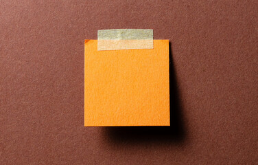 Piece of blank orange paper note with adhesive sticky tape and shadow on brown background