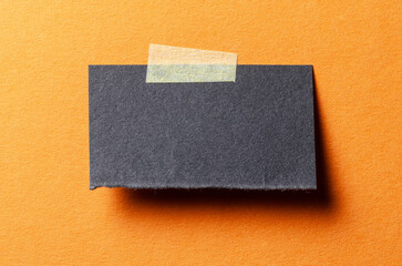 Piece of black paper note with adhesive sticky tape and shadow on orange background