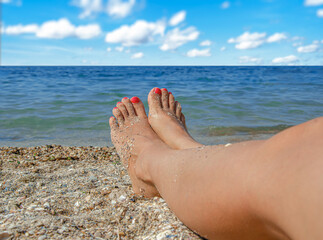 Sandy feet with red nail polish resting on a beach shore with a view of the sea and blue sky, representing relaxation and vacation lifestyle.