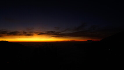 Fototapeta premium A brilliant orange and yellow glow from a sunrise or sunset illuminates the horizon over a dark, silhouetted landscape.