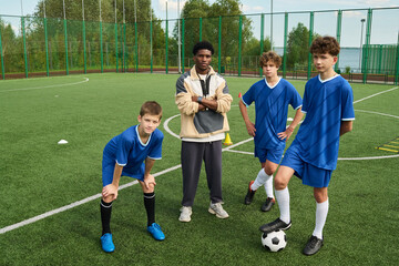 Obraz premium Black man standing with arms crossed coaching group of three teenage boys on outdoor soccer field, teenagers wearing uniforms and posing with soccer ball during training session