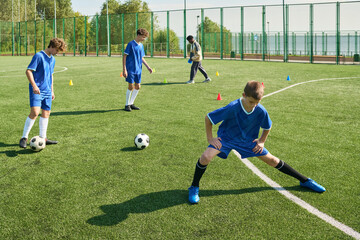 Three teenage boys practicing soccer drills on outdoor field while male coach observing in background, each boy focusing on ball control and footwork during training session