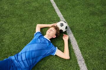 Teenage boy lying on grass soccer field resting with soccer ball under head eyes closed hands behind head wearing sports uniform relaxing after match © Mediaphotos