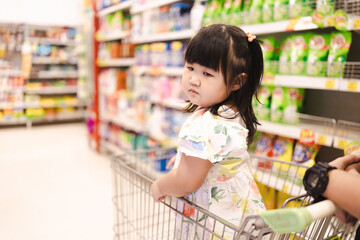A cute little Asian girl stands on a shopping cart while waiting for her mother to shop in a department store,happy infant baby sitting in shopping cart or trolley in grocery supermarket mother.