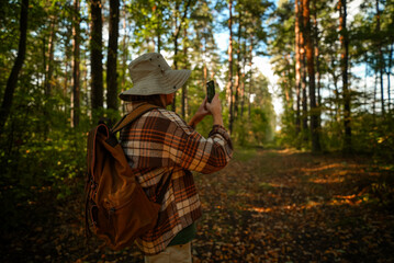 A tourist wearing a hat and carrying a backpack takes pictures of the forest on his phone